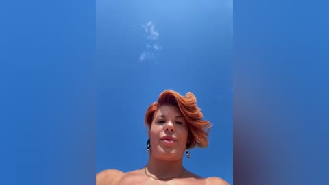 A video of a smiling, light-skinned woman with short, wavy red hair, wearing dark earrings, standing in front of a bright blue background with a faint cloud.