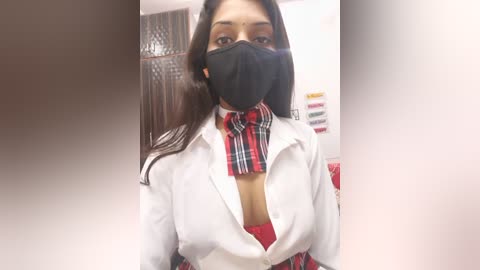 Video of a young woman with medium skin tone, wearing a black face mask, white button-up shirt, red plaid tie, and a red bra. Background shows a blurred, white-walled room with a bookshelf.