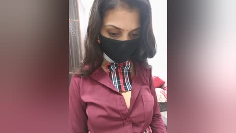 Video of a young woman with medium skin tone, wearing a black face mask, maroon button-up shirt, and a red and black plaid tie, standing in a dimly lit room with a blurred background.