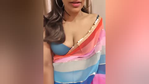 Video of a South Asian woman with medium skin tone, wearing a colorful, off-shoulder sari with blue, orange, pink, and white stripes, and large hoop earrings.