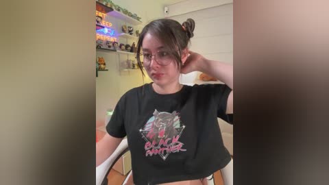 A video of a young woman with fair skin, dark hair in a bun, wearing glasses, a black \"Black Panther\" T-shirt, and a relaxed expression. Background includes shelves with toys, a neon sign, and a window.