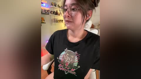 Video of a young woman with fair skin, glasses, and brown hair in a bun, wearing a black \"Batman\" T-shirt, seated in a gaming chair in a cozy room with shelves of toys and colorful LED lights.