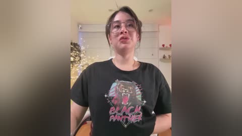 Video of a young woman with glasses and dark hair, wearing a \"Black Panther\" T-shirt, in a dimly lit room with Christmas decorations.