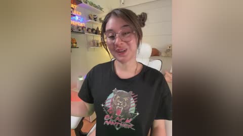 A video of a young woman with glasses, brown hair in buns, wearing a black graphic t-shirt with a lion design, in a cozy, brightly-lit room with shelves and colorful decorations.
