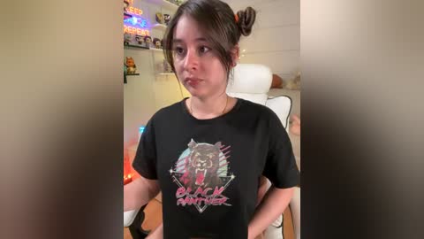 Media: Video of a young woman with dark hair tied back, wearing a black t-shirt with a pink and blue graphic design, seated in a white gaming chair, surrounded by gaming room decor.