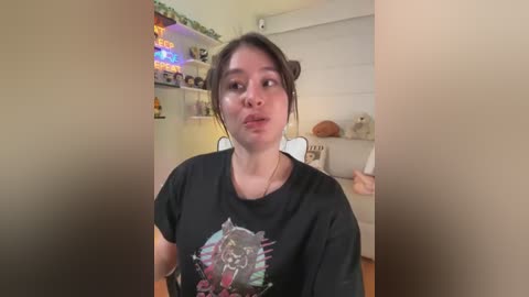 Video of a young woman with light skin and dark hair, wearing a black t-shirt with a colorful graphic design, in a cozy, dimly lit living room with shelves and plants.