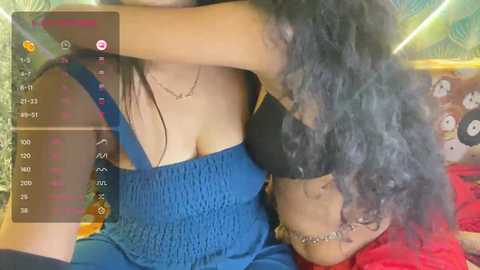Video of two women with long hair; one in a blue dress, the other in a black bra, kissing passionately in a colorful, playful room.