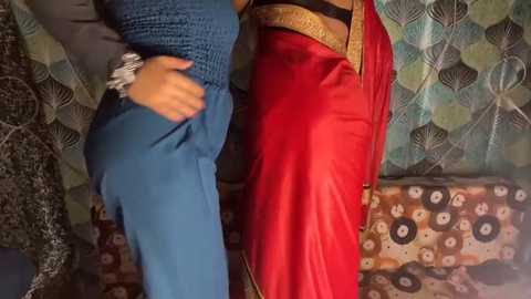 Video of two women in traditional attire, one in a blue knit top, the other in a vibrant red sari with gold trim, standing side by side in front of patterned curtains.
