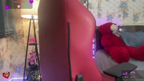 A video shows a cozy bedroom with a red leather bed frame, a plush red teddy bear, and a floral-patterned wallpaper. The room is dimly lit with a standing lamp and a pink phone charger.