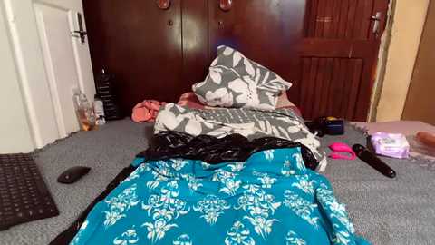 Video of a messy bedroom with a blue floral duvet, black pillow, and scattered clothes. A TV remote and a black keyboard are on the gray carpet, with a wooden door and a closet in the background.