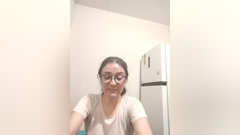 A video of a young woman with light skin and dark hair, wearing glasses, seated in a narrow, beige-walled room. She is wearing a beige shirt and looking forward. A white refrigerator is partially visible in the background.