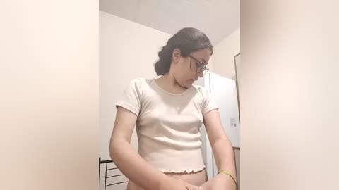 A video of a young, slender Asian woman with fair skin, wearing glasses and a white crop top, sitting on a metal chair in a beige-walled room.