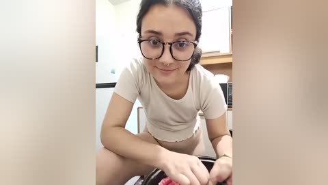 Video of a young woman with glasses and fair skin, wearing a short-sleeve beige crop top, sitting on a kitchen counter, washing dishes. The background shows a modern kitchen with white cabinets and a window.