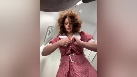 Video of a woman with curly hair in a red checkered dress, adjusting her bra, in a white-walled room with a gray curtain.