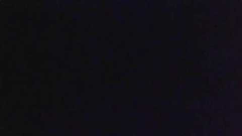 A dark, grainy video with indistinct, blurry figures in the background. The scene is mostly black, with faint, indiscernible shapes and a hint of purple and blue hues. The image is grainy, likely due to low light or a poor camera setting.