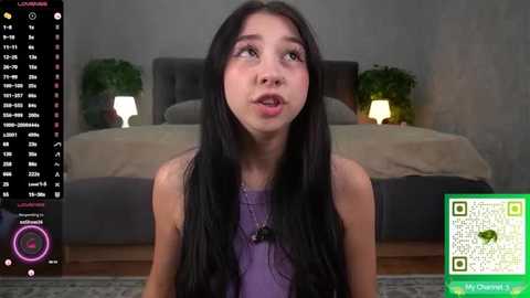 Video of a young Asian woman with long black hair, wearing a purple top, sitting in a modern bedroom with a grey bed, green plants, and a wooden floor.