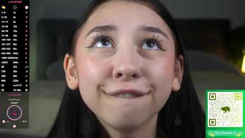 A close-up video of a young woman with fair skin, dark hair, and a nose ring, smiling. The background shows a digital screen displaying a calendar and a QR code.