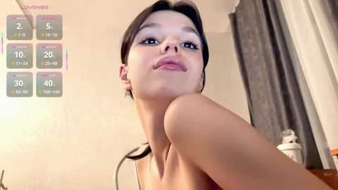 A video of a young, fair-skinned woman with dark hair and large eyes, wearing minimal makeup, taken indoors. The background shows a bathroom with a white sink, mirror, and window with grey curtains.