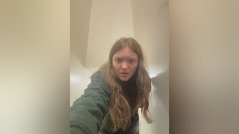 A video of a young woman with long, wavy brown hair, wearing a green jacket, taken from a low angle, creating a distorted perspective. She is standing in a beige room with a white door.