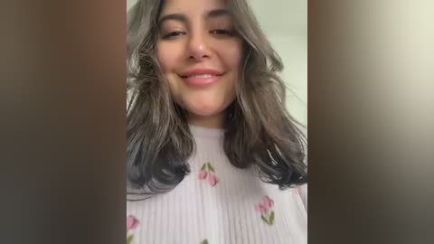 Video of a young woman with wavy, dark hair, fair skin, and a warm smile, wearing a white top with pink floral embroidery, set against a blurred background.