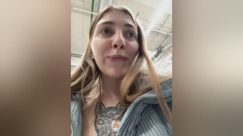 Video of a young Caucasian woman with light skin and long blonde hair, wearing a light blue, ribbed zip-up sweater, revealing a detailed, black-and-white tattoo of a woman's face on her chest. The background shows an industrial setting with metal structures and pipes.