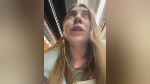 Media: Video of a young woman with long, blonde hair, wearing a dark top, looking up with a neutral expression. Background shows a beige wall and fluorescent lights.