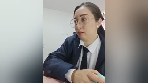 A video of a young Asian woman with glasses, wearing a navy blue blazer over a white shirt and black tie, sitting indoors, looking towards the camera.