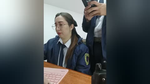 Video of a young woman with glasses, long brown hair, in a dark blue police uniform, sitting at a desk with a pink keyboard.