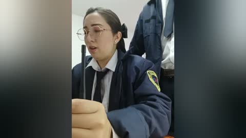 A video captures a young woman with fair skin, long dark hair, and glasses, dressed in a navy blue police uniform with a badge. She appears to be holding a small object, possibly a phone, while seated in a dimly lit room with blue jackets hanging nearby.