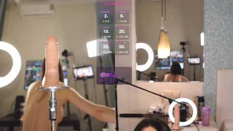 Video of a nude woman with a dildo on her head, surrounded by studio lights, in a modern, dimly lit room.