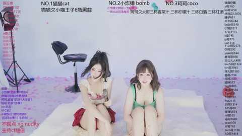 Video of two young Asian women with fair skin, sitting on a fluffy white rug. One wears a red skirt, the other a green bra. Background includes a stool and studio lighting equipment.