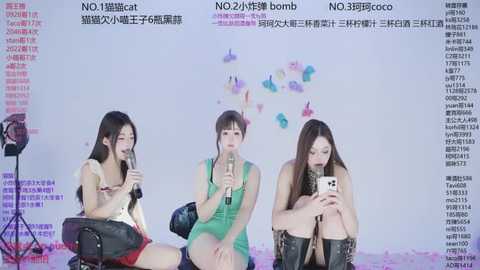 Video of three Asian women in colorful, sleeveless dresses, seated on a studio chair, singing into microphones against a white backdrop with butterfly decals, wearing black leather jackets.