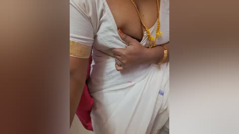 Video of a woman with medium brown skin wearing a white dress, holding her breast, adorned with gold jewelry and a red purse in the background.