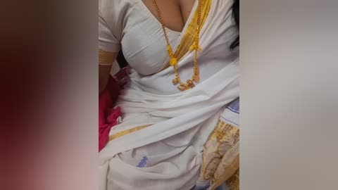 Video of a woman's torso in a white sari with gold embroidery, revealing cleavage, adorned with a gold necklace and red blouse peeking out.