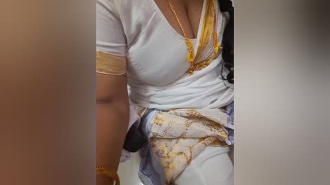 Video of a woman wearing a white sari with golden accents, a yellow blouse, and a gold necklace, partially obscured by a blurred figure in the foreground.