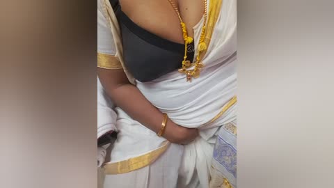 Video of a person in traditional South Asian attire, a white saree with gold trim, featuring a black bra underneath. The image focuses on the upper body, highlighting the intricate design and jewelry.