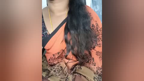 Video of a South Asian woman with medium brown skin, wearing an orange sari with black floral patterns. Her long, wavy black hair drapes over her shoulder. She has a gold necklace. Background is blurred.