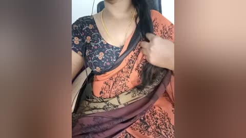 Video of a woman wearing an orange saree with floral patterns and a navy blue blouse, holding a beige sari, against a blurred background.