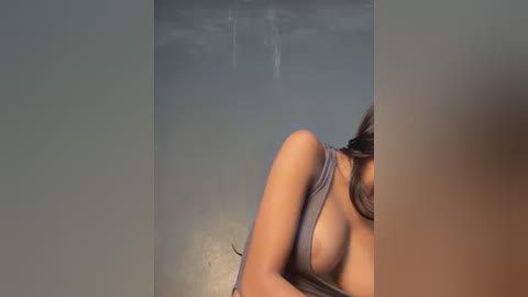 Video of a woman with medium skin tone, partially undressed in a grey tank top, exposing her breasts. Her long brown hair partially obscures her face. She is standing in a dimly lit room with a water-stained ceiling.