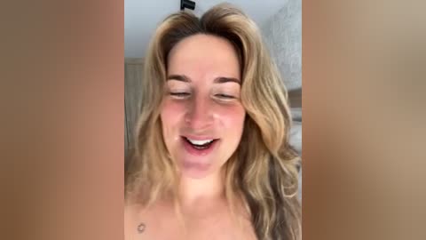 Video of a smiling, light-skinned woman with wavy blonde hair, wearing a small tattoo on her chest, standing indoors with blurred background.