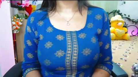 Video of a woman in a blue, patterned long-sleeve blouse with a square neckline. She wears a silver necklace. Background includes a plush teddy bear and floral decor.