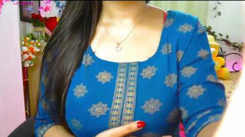 Video of a woman with fair skin and long black hair, wearing a blue kurta with gold embroidery and red nail polish. Background shows a colorful indoor setting with floral decorations and stuffed toys.