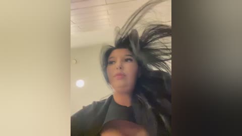 Video of a woman with long black hair, wearing a black top, captured mid-motion in a room with white walls and a ceiling light.