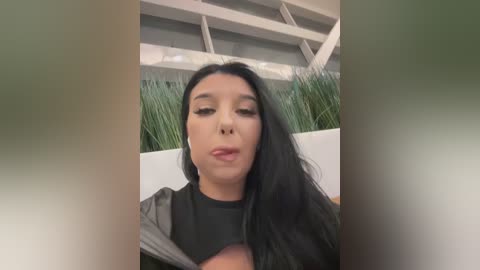 Video of a young woman with long black hair, closed eyes, and a neutral expression, wearing a black top, set against a modern indoor background with greenery and white beams.