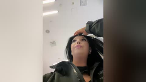 Video of a woman with long black hair, wearing a black jacket, adjusting her hair in a mirrored bathroom. The background shows white walls with ceiling lights.