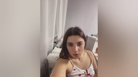 Video of a young woman with long brown hair, wearing a white tank top with red hearts, sitting on a gray couch in a cluttered, dimly lit room.