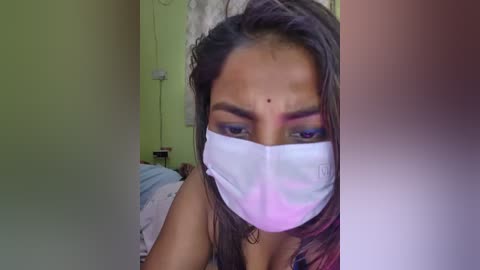 A video of a young South Asian woman with medium-brown skin, wearing a white surgical mask, brown hair, and a bindi on her forehead. She's indoors, partially visible in a blurred background.