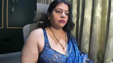 Video of a South Asian woman with medium skin tone, wearing a blue sequined saree, seated on a beige chair, in a room with green curtains and framed artwork.