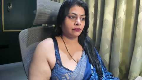 Video of a South Asian woman with medium skin tone and long black hair, wearing a blue sari and a gold necklace, seated in a grey upholstered chair, against a beige curtain background.