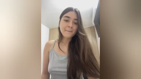 Video of a young woman with long, straight brown hair, light skin, and a slender physique, wearing a light gray tank top. She stands in a beige hallway with a wooden door and light-colored walls.